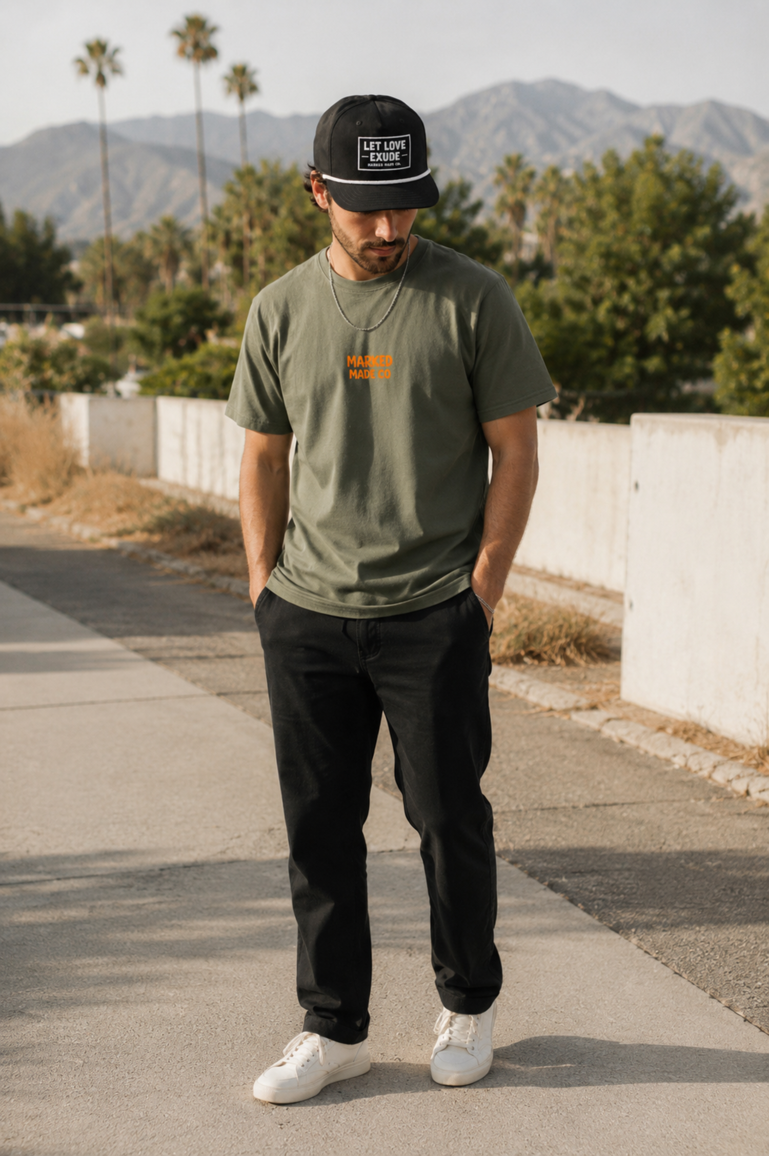 Man wearing a green t-shirt with orange text, black cap, and white sneakers standing on a sidewalk with mountains in the background.